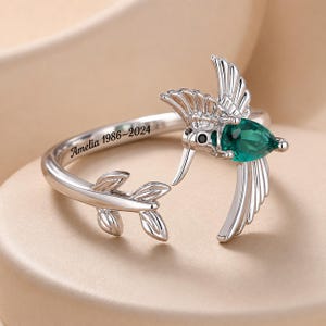 Puede incluir: Un anillo abierto de tono plateado con un diseño de colibrí, una piedra preciosa verde en forma de pera y un pequeño ojo de piedra negra. La banda está grabada con el texto Amelia 1986-2024 y termina en una pequeña rama con tres hojas.