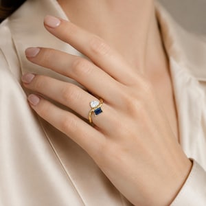 Puede incluir: Un anillo de oro con diseño bypass que presenta dos piedras preciosas distintas. Una piedra es un diamante transparente en forma de pera y la otra es un zafiro azul profundo de corte rectangular. El aro está hecho de oro amarillo pulido.