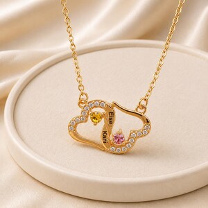 Puede incluir: Un collar de tono dorado con un colgante de dos corazones entrelazados. El colgante está adornado con piedras de circonita cúbica transparentes, una piedra amarilla y una piedra rosa. Los nombres Eise y Kani están grabados en el centro.