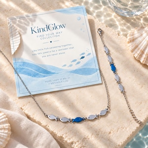 May include: A silver-toned necklace and bracelet set featuring small fish-shaped charms, with one blue fish charm on each piece. The jewellery is displayed with a square card printed with the text KindGlow Find Your Way Collection.