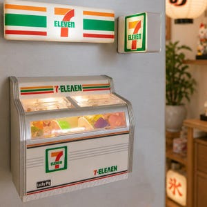 May include: A 7-Eleven themed wall-mounted display case featuring a silver frame, clear front panels, and the 7-Eleven logo. Above it are two illuminated 7-Eleven signs, one rectangular and one square, displaying the brand name and logo.