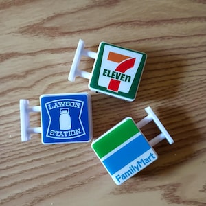Może przedstawiać: Trzy miniaturowe kwadratowe plastikowe szyldy z logo 7-Eleven, Lawson Station i FamilyMart. Każdy szyld jest biały i ma odrębną kolorystykę: zielony, pomarańczowy i czerwony dla 7-Eleven, niebieski i biały dla Lawson oraz zielony i niebieski dla FamilyMart.