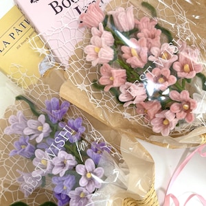 May include: Two bouquets of felt flowers in pink and purple, wrapped in brown paper with white lace netting and clear plastic film. The plastic film is printed with the text BEST WISHES for you. The flowers have green stems and yellow centers.