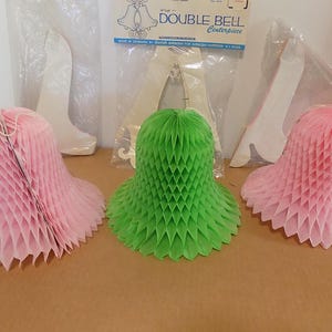 May include: Three honeycomb paper bell decorations in pink and green. The bells are in a bell shape with a ruffled edge. A package of a Double Bell centerpiece is in the background.
