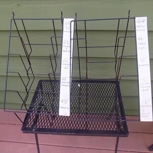 May include: A black metal display rack with multiple shelves and a diamond-patterned base. Two white signs with handwritten text are attached to the rack. The rack is on a wooden surface with a green background.