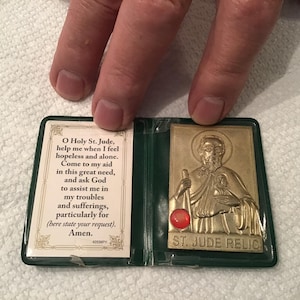 May include: A gold-colored St. Jude relic in a green plastic case. The relic features an image of St. Jude and the text "ST. JUDE RELIC". The case also contains a prayer card with text.