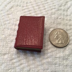 May include: A miniature burgundy book with a textured cover, next to a U.S. quarter for size comparison. The book's spine has raised detailing. The quarter shows the profile of George Washington.