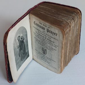 May include: An open, antique book with a burgundy leather cover. The title "The Book of Common Prayer" is visible on the right page. A black and white illustration is on the left page. The book's pages are aged and yellowed.