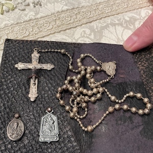 May include: A silver-toned rosary featuring a detailed crucifix and spherical beads, accompanied by two small religious pendants. One pendant is oval-shaped with a figure, and the other is shield-shaped with an embossed scene, all resting on a dark textured surface.