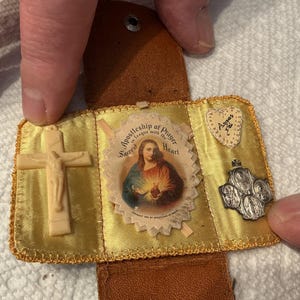May include: A vintage religious prayer case with a cream-colored crucifix, a small image of Jesus, and a silver medal. The case is made of gold satin fabric and brown leather. The text "Apostleship of Prayer" is visible.