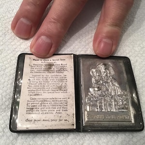 May include: A small, black, leather-bound prayer book with a silver image of Saint Anne and the Blessed Virgin Mary on the front cover. The book has a white paper insert with a prayer to Saint Anne.