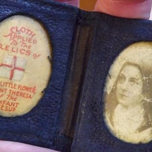 May include: A small, open, dark blue leather case with two oval inserts. One insert features a portrait of a woman, the other has text that reads "CLOTH APPLIED TO THE RELICS of THE LITTLE FLOWER SAINT THERESA OF THE INFANT JESUS" with a cross.