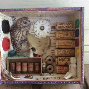 May include: A small wooden box filled with various objects, including a clock face, a small porcelain doll, a cork with the text "JEAN PANIS VIGNERON", and a printed image of an owl. The box is decorated with a purple and green border.