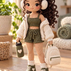 May include: A crocheted doll with long curly brown hair, wearing white earmuffs, a cream-colored zip-up jacket, an olive green crop top, and matching drawstring shorts. The doll wears white socks and olive green sneakers with white heart details.