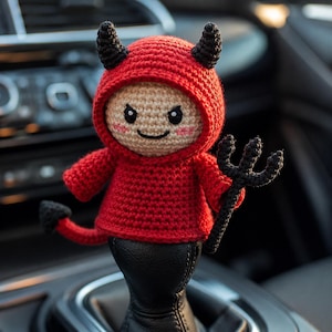 May include: A small, handmade crochet devil doll featuring a bright red hooded sweater, black horns, and a matching black pitchfork. The doll has a beige face with embroidered black eyes, a thin smile, and pink cheeks, plus a small red tail.