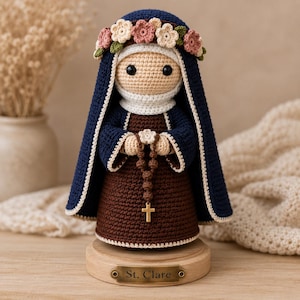 May include: A crocheted doll of St. Clare wearing a dark blue habit with a white trim, a brown dress, and a floral crown. The doll holds a brown beaded rosary with a gold cross. It stands on a round wooden base with a metal plaque reading St. Clare.
