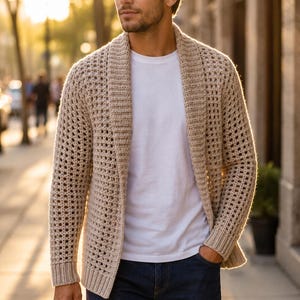 Può includere: Un cardigan beige aperto sul davanti con un motivo a uncinetto testurizzato a griglia, un ampio collo a scialle e polsini a coste. Il capo è indossato sopra una t-shirt bianca semplice e un paio di jeans in denim blu scuro.