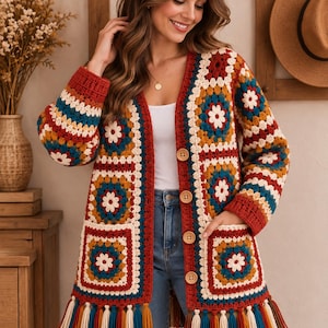 Op de afbeelding: Een gehaakt vest met lange mouwen en een veelkleurig granny square-patroon in roest, teal, mosterdgeel en crème. Het kledingstuk heeft een V-hals, vier grote houten knopen aan de voorzijde, twee opgestikte zakken en een afwerking met franjes.