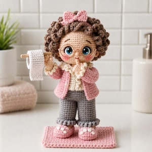 Puede incluir: Una muñeca de ganchillo con pelo castaño rizado, un lazo rosa y gafas redondas. La muñeca lleva una rebeca rosa sobre una camisa blanca con volantes y pantalones grises, con zapatillas rosas. Sostiene un pequeño rollo de papel higiénico tejido.