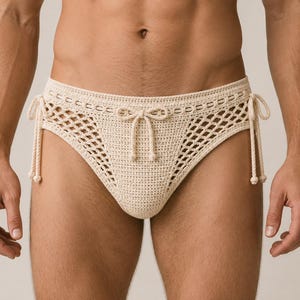 May include: A cream-colored crochet bikini bottom featuring a solid front panel, open-knit side panels, and adjustable side ties with a decorative bow at the center front. The garment is crafted from a textured yarn with a consistent stitch pattern.