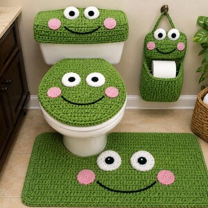 May include: A green crochet bathroom set featuring a frog motif. The set includes a rectangular tank cover, a round toilet seat cover, a rectangular floor mat, and a hanging toilet paper holder, all decorated with white eyes and pink cheeks.