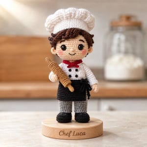 May include: A crochet doll of a chef wearing a white hat, white jacket with black buttons, a red necktie, and a black apron. The doll has brown hair, rosy cheeks, and holds a brown rolling pin. It stands on a round wooden base engraved with Chef Luca.