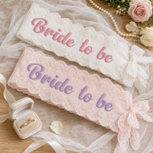 Puede incluir: Dos bandas de encaje con bordes festoneados, una blanca con texto bordado en rosa Bride to be y una rosa claro con texto bordado en morado Bride to be, cada una con un detalle de lazo de encaje en el extremo de la banda.