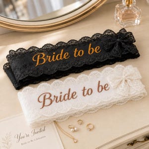 Puede incluir: Dos bandas de encaje, una negra y otra blanca, cada una con el texto bordado "Bride to be" en letra cursiva y un lazo de encaje decorativo en el lado derecho. Las bandas tienen bordes festoneados y se muestran junto a joyas y una invitación.