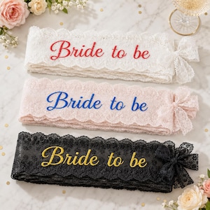 Puede incluir: Tres bandas de encaje con bordes festoneados y un lazo lateral. La banda superior es blanca con texto bordado en rojo Bride to be. La banda central es rosa claro con texto bordado en azul Bride to be. La banda inferior es negra con texto bordado en dorado Bride to be.