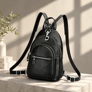 May include: A black leather backpack with a pebbled texture, featuring a rounded top, a front zipped pocket, and silver-toned metal hardware. The bag includes adjustable shoulder straps attached with a central silver ring and a top handle.