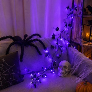 Op de afbeelding: Een decoratieve rankenslinger met donkere bladeren en kleine, gloeiende paarse LED-lampjes. De slinger is gedrapeerd over een lichtgekleurd oppervlak naast een grote, pluizige zwarte spin, een realistische menselijke schedel en een kussen met spinnenwebpatroon.