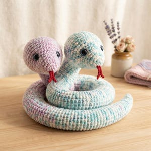 Può includere: Un serpente di peluche all'uncinetto a due teste, con una testa in varie tonalità pastello viola e l'altra in varie tonalità pastello blu e bianco. Entrambe le teste hanno occhi in plastica scura e una piccola lingua rossa all'uncinetto.