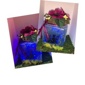 May include: A glass jar with a fairy silhouette inside, surrounded by blue lights. The jar is decorated with moss and a red flower on top.
