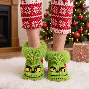 May include: A pair of bright green crochet slippers featuring a character face with yellow eyes, a black stitched smile, and green faux fur trim at the top. The slippers are worn with red and white patterned leggings featuring reindeer and snowflakes.