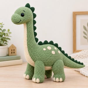May include: A crocheted dinosaur toy in shades of sage green and cream. The creature features a long neck, a row of dark green spikes along its back, and cream-colored spots on its side. It has black safety eyes and a stitched smile.