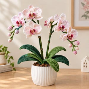 Könnte beinhalten: Eine gehäkelte Orchidee mit hellrosa und weißen Blütenblättern, dunkelrosa Zentren und grünen Blättern, eingetopft in einen weißen, gerippten Keramiktopf. Die Pflanze hat zwei Stängel mit mehreren blühenden Blüten und kleinen Knospen.