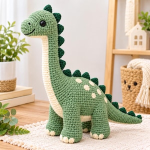 May include: A crochet dinosaur toy featuring a long neck and tail, crafted in sage green yarn with a cream-coloured belly and underside. It has dark green spikes along its back, cream-coloured spots on its side, and large black safety eyes.
