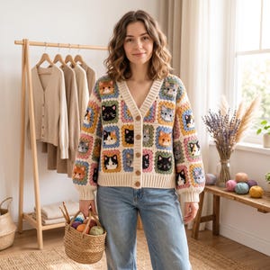May include: A V-neck cardigan made of crocheted granny squares, each featuring a different cat face in various colors including orange, black, white, and gray. The sweater has long sleeves, a cream-colored ribbed trim, and four brown buttons down the front.