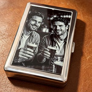 May include: A rectangular silver metal case featuring a black and white engraved image on the front. The image depicts two individuals smiling while holding glasses of beer. The case has a polished, reflective metallic finish and a hinged lid.