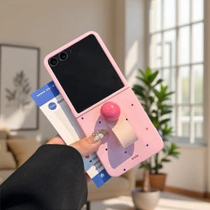 May include: A light pink foldable smartphone case featuring a dark screen, a circular pink knob, and a light pink strap. The case is decorated with small colorful dots and the word smile printed in black. A blue and white card is tucked behind the case.