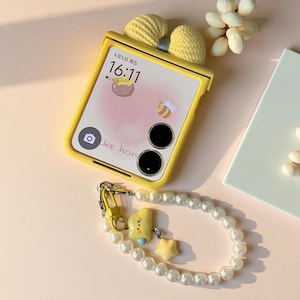 May include: A yellow foldable smartphone case featuring a textured yellow bow at the top. The screen displays the time 16:11, a bee icon, and a honey pot graphic. A detachable wrist strap made of white faux pearls with a yellow cloud and star charm.