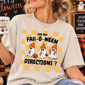 May include: A beige t-shirt featuring a yellow checkered background with three white ghosts wearing orange beanies and holding orange drinks. The text reads ARE YOU FALL-O-WEEN DIRECTIONS? in black letters. The ghosts are also holding pumpkins.