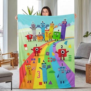 May include: A soft fleece blanket featuring a colorful Numberblocks graphic print. The design displays various numbered block characters in red, blue, yellow, green, and purple, arranged over a rainbow path against a light blue sky background.