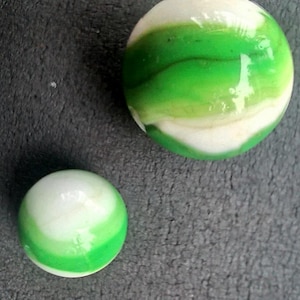 May include: Two spherical glass marbles of different sizes, each featuring a smooth, glossy surface with swirling patterns of opaque white and vibrant green colors that wrap around the round bodies of the objects in a continuous, flowing design.