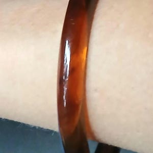 May include: A smooth, circular bangle bracelet with a translucent, deep amber-brown color. The material has a glossy, polished finish that catches the light, showing subtle variations in tone throughout the solid, rounded band structure.
