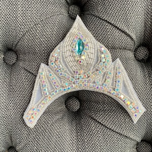 May include: A silver and white crown with a blue jewel in the center. The crown is made of beads and sequins.