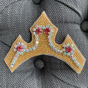 Può includere: Una corona dorata con strass rosa e trasparenti. La corona è fatta di perline ed è cucita su un tessuto bianco.