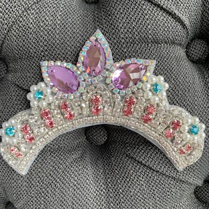 May include: A silver tiara with a curved base, adorned with pink and blue gemstones, white pearls, and large purple teardrop-shaped gems. The tiara is embellished with clear rhinestones and is set against a gray background.