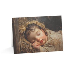 May include: A greeting card featuring a painting of a sleeping infant with dark curly hair resting on a bed of dry golden straw. The infant is wrapped in a light beige fabric, with one hand tucked near the face, set against a dark background.