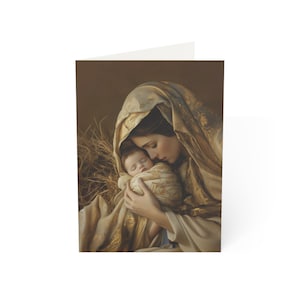 May include: A greeting card featuring a painting of a figure in a gold-toned head covering holding a swaddled infant. The pair is nestled against a background of dry straw, rendered in soft, warm brown and golden hues with detailed fabric textures.